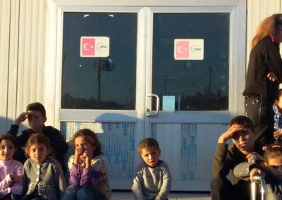 Na zdjęciu kilkuletnie dzieci siedzące na schodach przed drzwiami do kontenera. Na nich naklejki organizacji Czerwonego Półksiężyca, nad nimi napis Midjat i inne w języku tureckim i arabskim oraz logo junisefu. Po prawej stronie kobieta oraz chłopiec z dziewczynką na wózku inwalidzkim.