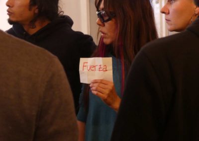 Na zdjęciu brunetka z grzywką w okularach z grubymi oprawkami, trzymająca w dłoni karteczkę z czerwonym napisem Fłerza. Jest otoczona kilkoma osobami, na zdjęciu tylko ich fragmenty. Spoglądają w lewą stronę.