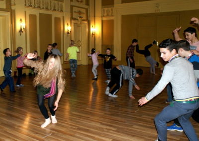 Zdjęcie przedstawia młodzież i dzieci na tanecznej zabawie w sali o skromnym neobarokowym stylu. Niektórzy wykonują skomplikowane pozy, jak stanie na rękach, inni tańczą spokojniej.