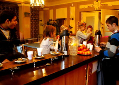 Zdjęcie przedstawia dzieci i młodzież przy barze. Na nim termosy z napojami, dzbany z lemoniadą i owoce. Bufet znajduje się w sali o stylistyce lat siedemdziesiątych.