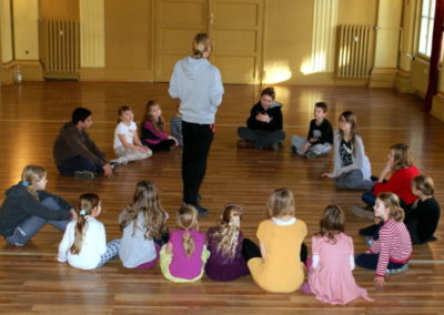 Na zdjęciu dzieci w wieku początkowoszkolnym siedzą w okręgu na podłodze. W jego środku stoi odwrócony chłopak w jasnych, spiętych w kitkę włosach i szarej bluzie.