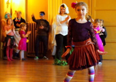 Na zdjęciu tańcząca dziewczynka w brązowej rozkloszowanej spódniczce i pasiastych rajstopkach. Za nią stoją inne dzieci, niektóre poprzebierane.