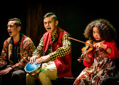 Na zdjęciu dwóch nastolatków śpiewających i grających na bębnach oraz ciemnoskóra dziewczynka w bujnych kręconych włosach grająca na skrzypcach. Chłopak po lewej stronie ma kurteczkę w etniczne wzory, siedzący w środku czerwoną kamizelkę i kraciastą koszulę, patrzy w obiektyw. Dziewczynka w kwiecistej sukience skupia się na grze.
