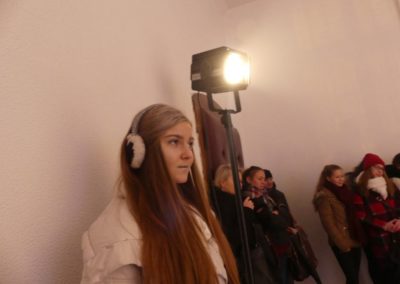 Na zdjęciu stojąca przy reflektorze scenicznym okrągła nastolatka w długich prostych włosach. Na uszach ma futrzane słuchawki. Spogląda przed siebie, w prawą stronę. Za nią stoi kilka osób w kurtkach.