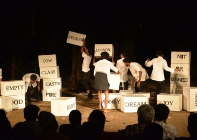 Na scenie siedem osób ubranych na biało czarno. Krzątają się wśród białych pudeł z angielskimi napisami. Układają jedne na drugie. Przed nimi głowy publiczności.