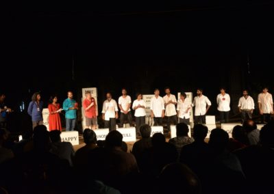 Na scenie grupa kilkunastu osób, w większości hinduskiej urody. po lewej stronie ubrani na kolorowo, po prawej na biało czarno. Stoją za białymi pudłami z angielskimi napisami. Mężczyzna wypowiada się przez mikrofon. Przed nimi siedząca publiczność.