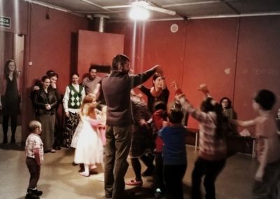 Na zdjęciu grupa dorosłych i dzieci. Część z nich tańczy. Są w wymalowanej na czerwono sali.
