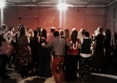 Na nieco niewyraźnym zdjęciu grupa ludzi w wymalowanej na czerwono sali oświetlonej jadowitymi żarówkami. Niektórzy tańczą, inni stoją. Są odwróceni tyłem do obiektywu.