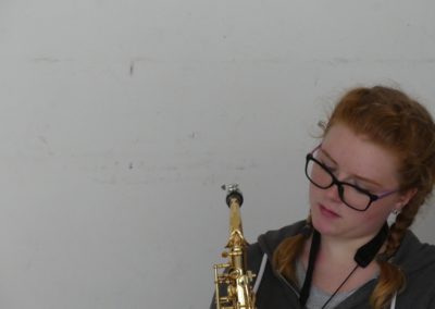 Na zdjęciu ubrana w szarości ruda nastolatka w okularach. W dłoniach trzyma złoty instrument dęty. Stoi przed białą ścianą.