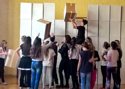 Na zdjęciu grupa młodzieży układająca wysoką ścianę z pudełek. Jeden z chłopców siedzi na czyichś ramionach i stawia kartony w najwyższym rzędzie. Znajdują się w sali o miodowych ścianach.