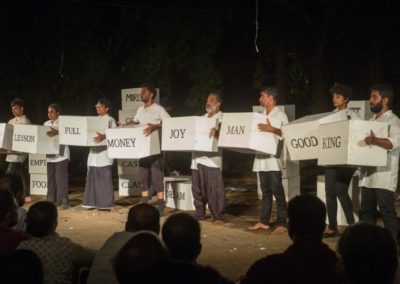 Na zdjęciu grupa ośmiu mężczyzn urody hinduskiej trzymających w dłoniach przed sobą białe pudła z napisami w języku angielskim. Wszyscy są ubrani w białe długie koszule i rozmaite czarne spodnie. Przed nimi siedząca publiczność. Za nimi owity ciemnością las.