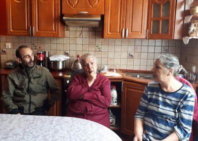 Na zdjęciu dwie siwiejące kobiety i mężczyzna w wieku przedemerytalnym. Siedzą przy stole, za nimi kuchenne meble. Są ubrani skromnie. Najstarsza kobieta, siedząca w środku się wypowiada, pozostali na nią spoglądają.