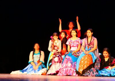 Zdjęcie przedstawia sześć dziewczynek w tradycyjnych, kolorowych sukniach romskich. Siedzą na ławie i ziemi. Wszystkie są brunetkami, uczesanymi w warkocze. Z tyłu kobieta w średnim wieku w czerwonej bluzce, ma uniesione dłonie.