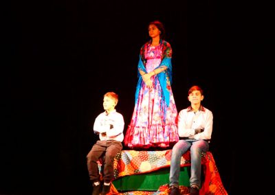 Zdjęcie przedstawia dziewczynkę w tradycyjnym, kolorowym stroju romskim i narzuconej na ramiona barwnej chuście. Stoi na wyłożonym materiałami podeście. Poniżej siedzi dwóch chłopców ubranych w białe koszule. Są w ciemnej sali.