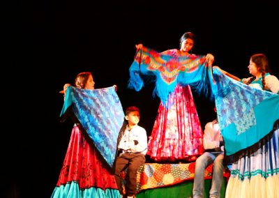 Zdjęcie przedstawia trzy dziewczynki w tradycyjnych, kolorowych sukniach romskich. W dłoni trzymają rozłożone barwne chusty. Stoją na podwyższeniu pokrytym materiałami. Poniżej siedzi dwóch chłopców w białych koszulach. Są w ciemnej sali.