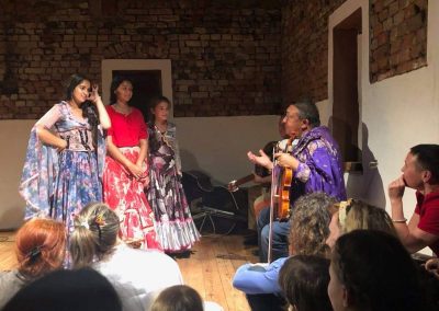 Na zdjęciu trzy dziewczynki w kwiecistych romskich sukienkach, przysłuchują się wypowiedzi starszego mężczyzny ze skrzypcami w dłoni. Przed nimi skupiona publiczność, za nimi dwie pary drzwi i ceglana ściana.