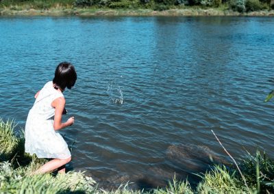 Na zdjęciu brunetka we włosach do ramion puszczająca kaczki, czyli rzucająca kamienie do rzeki. stoi na jej brzegu, tyłem do nas odwrócona. Jest w jasnej, letniej sukience, jest słoneczny dzień.