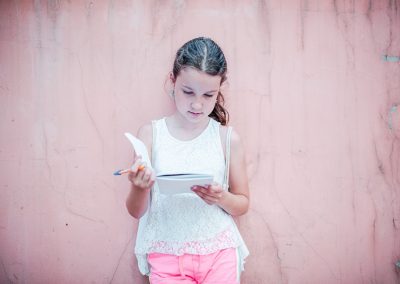 Na portretowym zdjęciu zamyślona dziewczynka przyglądająca się otwartemu zeszytowi, który trzyma w dłoniach. Ma ciemne włosy, jest w białej koszulce i różowych spodenkach. Stoi oparta o różową, nieco przybrudzoną ścianę.