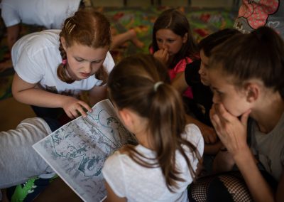 Na zdjęciu pięć dziewczynek przyglądających się niewyraźnej mapie z zaznaczeniami. Trzyma ją szatynka uczesana w kitkę. Siedzą na wykładzinie z dziecięcym widokiem miasta.