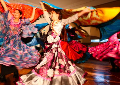 Zdjęcie przedstawia wirujące w tańcu na scenie dziewczynki w barwnych tradycyjnych romskich sukniach. W dłoniach trzymają rozciągnięte za sobą kolorowe chusty.