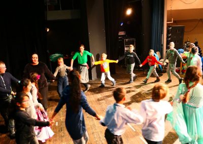 Zdjęcie przedstawia tańczące w okręgu dzieci w różnym wieku. Trzymają się za ręce, większość jest rozradowana.
