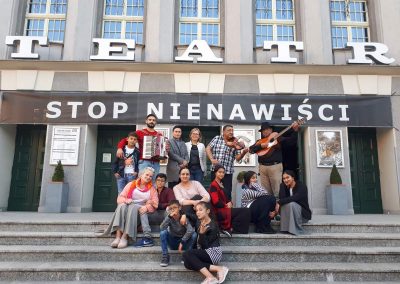 Na zdjęciu grupa ludzi przed budynkiem z napisami: Teatr, i stop nienawiści. Niektórzy siedzą na schodach, inni stoją. Mężczyźni w ostatnim rzędzie mają instrumenty. Gitarę, akordeon i gitarę.