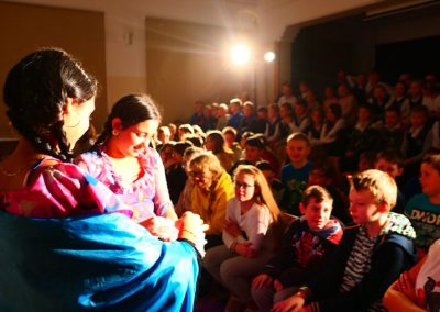 Zdjęcie przedstawia dwie Romki, które wyszły w trakcie spektaklu do publiczności. Są w warkoczach i kolorowych sukienkach. Większość osób z publiczności to dzieci.