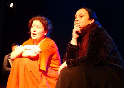 Na zdjęciu dwie brunetki w średnim wieku. Siedzą z podkurczonymi kolanami. Po lewej wypowiadająca się kobieta w pomarańczowej sukni, po prawej ubrana w brązy zamyślona kobieta.