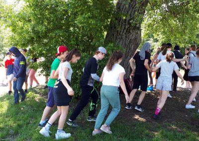 Na zdjęciu grupa młodzieży okrążająca w parach drzewo. Trzymają się za ręce. Są w letnich, sportowych ubraniach, wglądają na zmęczonych.
