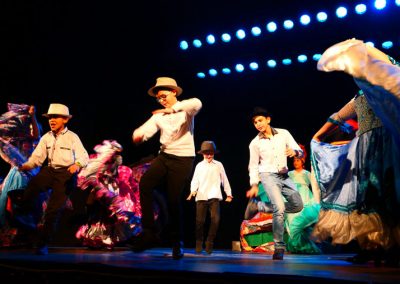 Zdjęcie przedstawia fragment spektaklu Baśń Żywa Fundacji Art Dżankszon. Tańczący chłopcy w białych koszulach i kapeluszach oraz dziewczynki w barwnych romskich sukniach, które wirują w tańcu. Z boku mężczyzna grający na akordeonie.