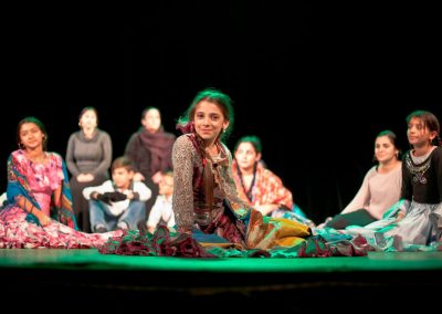 Zdjęcie przedstawia dziewczynkę, dziewczyny i kobiety romskie siedzące bokiem na wyłożonej na zielono scenie. Wszystkie są ubrane w długie spódnice.