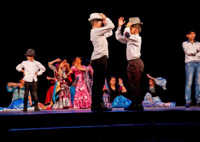 Zdjęcie przedstawia fragment spektaklu Baśń Żywa Fundacji Art Dżankszon. Na przedzie tańczący chłopcy w białych koszulach i kapeluszach. Za nimi tańczą i siedzą dziewczynki w barwnych tradycyjnych romskich sukniach.