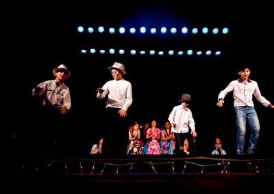 Zdjęcie przedstawia fragment spektaklu Baśń Żywa Fundacji Art Dżankszon. Na przedzie tańczący chłopcy w białych koszulach i kapeluszach. Za nimi siedzą dziewczynki w barwnych tradycyjnych romskich sukniach. Nad nimi dwa rzędy białoniebieskich reflektorów.