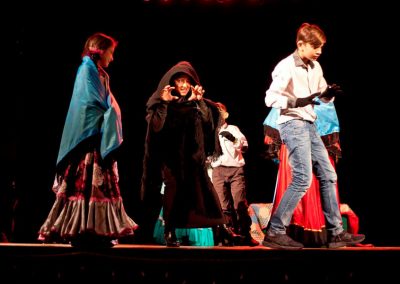 Zdjęcie przedstawia fragment spektaklu Baśń Żywa Fundacji Art Dżankszon. Na scenie dziewczynka w romskiej sukni z narzuconą niebieską peleryną, chłopiec w białej koszuli i kobieta w brązach i kapturze grająca groźną wiedźmę lub wilka. Za nimi inne postacie.
