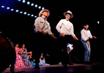 Zdjęcie przedstawia fragment spektaklu Baśń Żywa Fundacji Art Dżankszon. Na przedzie tańczący chłopcy w białych koszulach i kapeluszach. Za nimi siedzą dziewczynki w barwnych tradycyjnych romskich sukniach.
