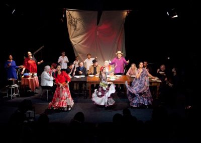 Na środku sceny tańczą trzy dziewczyny w długich romskich sukniach z falbanami. Za nimi ludzie siedzący przy zastawionym drewnianym stole. Po bokach muzykujące osoby, a na dole zdjęcia cienie publiczności.