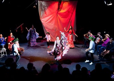 Na scenie grupa w romskich strojach. Na środku trzy dziewczyny w długich sukniach z falbanami, oraz trzech tańczących chłopców w kapeluszach z wiankami. Po bokach muzykujące osoby, a na dole zdjęcia cienie publiczności.