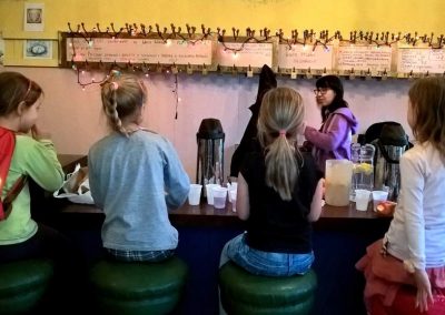 Dziewczynki siedzą na krzesłach barowych przy barowej ladzie, zastawionej termosami, dzbankami i kubeczkami z napojami. Za barem młoda kobieta w okularach, czarnych włosach i fioletowej bluzie stoi pod dawnymi wieszakami szatniowymi, teraz ozdobionymi lampkami choinkowymi.