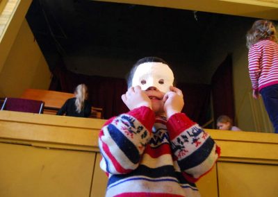 Mały chłopiec w pasiastym sweterku z reniferami zasłania twarz wykonaną najprawdopodobniej z gipsu prostą maseczką. Stoi przed sceną, na niej krzątają się dziewczynki.