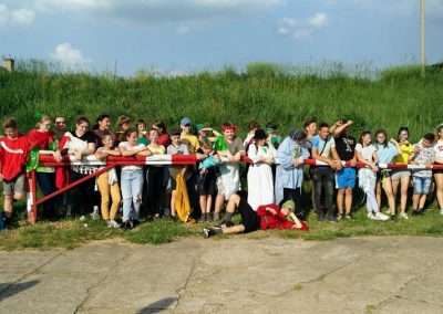 Zdjęcie przedstawia dużą grupę dzieci i młodzieży opierających się o biało czerwony szlaban. Tuż za nimi zielona łąka na pagórku.