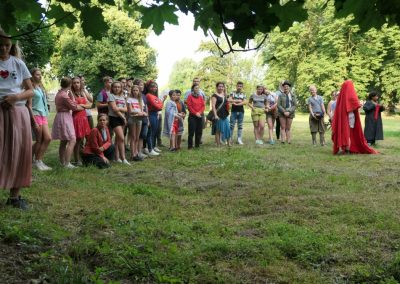 Na zdjęciu duża grupa nastolatków stojących w półokręgu na trawniku w parku. Są ubrani w swobodne stroje. wyjątek stanowi postać na środku, która jest okryta, wraz z twarzą, czerwonym materiałem, niczym mnich. Wokół są drzewa, jest słoneczny, letni dzień.