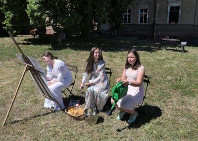 Na zdjęciu trzy nastolatki siedzące na krzesełkach na trawniku przed dworkiem w Mikuszewie. Jedna z nich rysuje na sztaludze, siedząca w środku obiera ziemniaki, siedząca zaś po prawej podlewa trawę z zielonej konewki.
