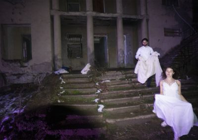 Zdjęcie przedstawia ubranych na biało parę młodych ludzi zbiegających po schodach prowadzących do zrujnowanego budynku. Dziewczyna jest w białej letniej sukni, choć na ziemi i schodach są ślady śniegu.