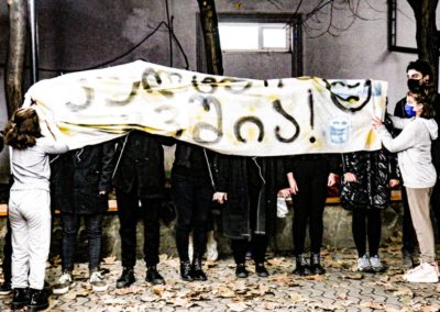 Zdjęcie przedstawia grupę młodych ludzi ubranych na czarno, których twarze są zasłonięte dużym bannerem z napisami w języku i alfabecie gruzińskim na białym materiale. Banner przytrzymują dwie dziewczyny ubrane na biało. Stoją na zewnątrz, za nimi okno, na ziemi opadłe liście.