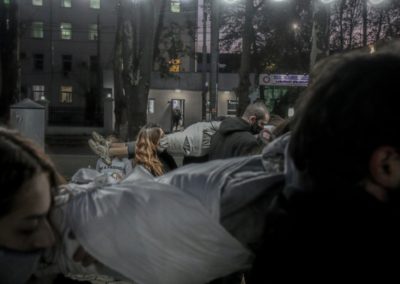 Zdjęcie przedstawia stojącą na ulicy grupę smutnych ludzi w maseczkach ochronnych na twarzy. Na ramionach trzymają owinięte w płachtę materiału ciało chłopaka. Wszyscy są performerami podczas pokazu Miasto jako inspiracja na ulicach Gori w Gruzji.