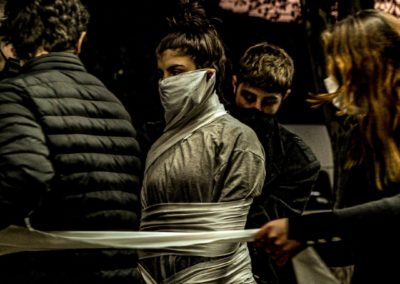 Zdjęcie przedstawia stojącą na ulicy grupę ludzi w maseczkach ochronnych na twarzy. Wśród nich dziewczyna w koku, która jest owinięta w płachtę materiału. Wszyscy są performerami podczas pokazu Miasto jako inspiracja na ulicach Gori w Gruzji.