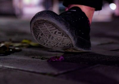Zdjęcie przedstawia zbliżenie podeszwy sportowego, czarnego buta, tuż przed zdepnięciem leżącego na chodniku zerwanego, fioletowego kwiatka.