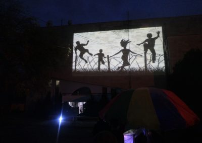 Zdjęcie przedstawia wyświetlaną z rzutnika na ścianie dużego budynku grafikę projektu Art łidałt border Fundacji Art Dżankszon. Na niej czarne cienie trzech chłopców i kobiety przeskakujących radośnie przez płot z drutu z zasiekami.