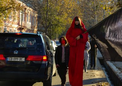 Zdjęcie przedstawia dziewczynę w czerwonej długiej pelerynie na szczudłach. Idzie ulicą, wśród samochodów. Niesie ogromną czarną płachtę.