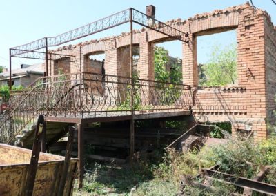 Zdjęcie przedstawia ruinę dawnego ceglanego budynku mieszkalnego z ażurowym metalowym gankiem charakterystycznego dla Gruzji i schodami. Przed budynkiem zardzewiałe żelastwa. Jest słoneczny dzień.